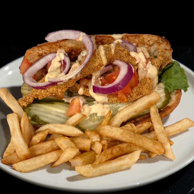 Crispy fried fish po'boy sandwich with red onions, tomatoes, pickles, and fries on a white plate.